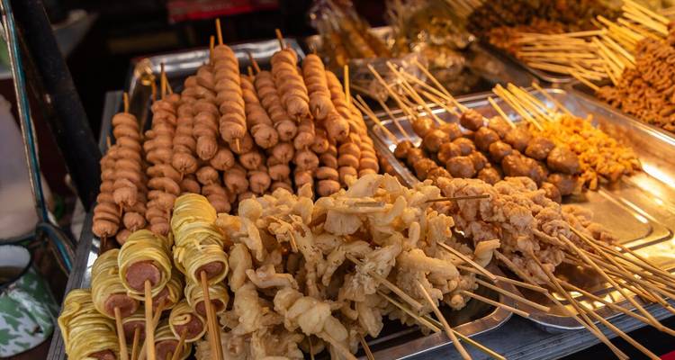 Primer plano de varios bocadillos indonesios pinchados cuidadosamente organizados en bandejas de metal en un mercado nocturno.