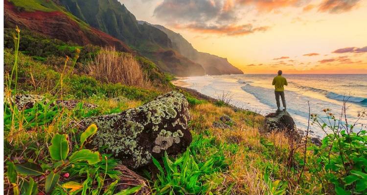 Person standing on a rocky shoreline at sunset with a colorful sky.