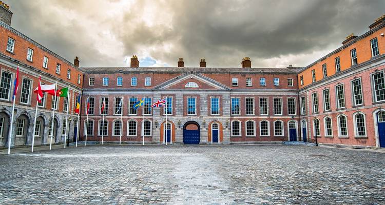 La cour imposante en briques rouges du château de Dublin est encadrée par des colonnades en arches sous des nuages dramatiques.