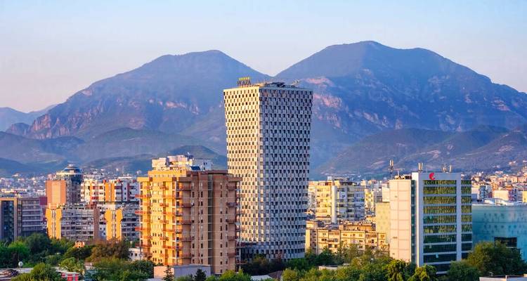 Stadsgezicht met hoge gebouwen en bergen op de achtergrond tijdens zonsondergang.