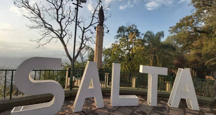 Grote witte SALTA letters die uitkijken over een schilderachtig uitzichtpunt omlijst door bomen en een verre obelisk.