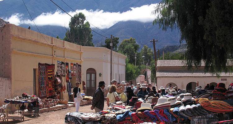 Drukke kunstnijverheidsmarkt met toeristen die kleurrijke textiel bekijken tegen een bergachtige achtergrond.