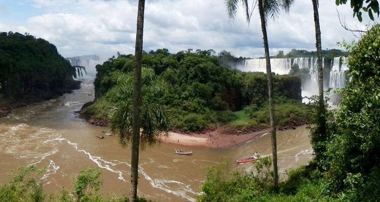 Panoramisch uitzicht op de Iguazu-rivier die zich splitst rond een weelderig eiland met boten nabij modderige oevers.