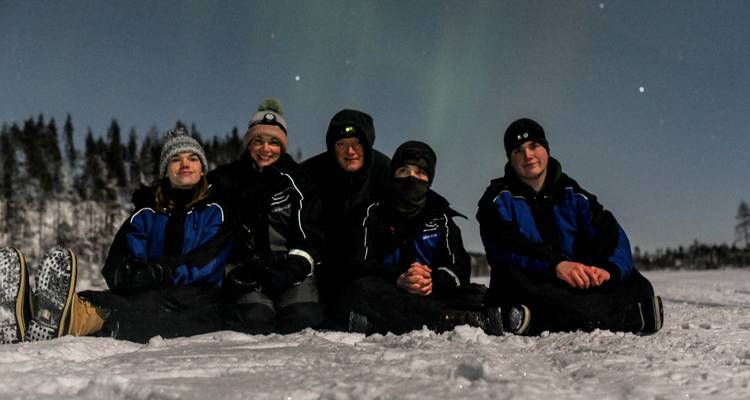 Cinco amigos en trajes de nieve se sientan en un lago congelado bajo tenues auroras boreales verdes y un cielo estrellado.