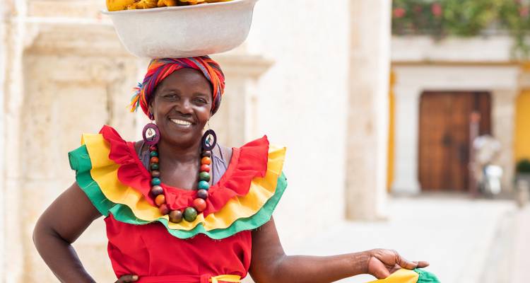 Femme en tenue colorée équilibrant un panier de fruits sur sa tête.