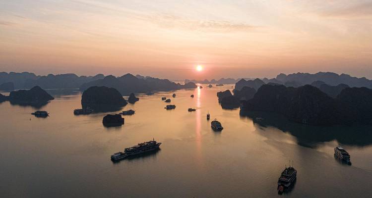 Panorama aérien de la baie d'Halong au lever du soleil avec des dizaines de bateaux de croisière dispersés sur des eaux calmes.