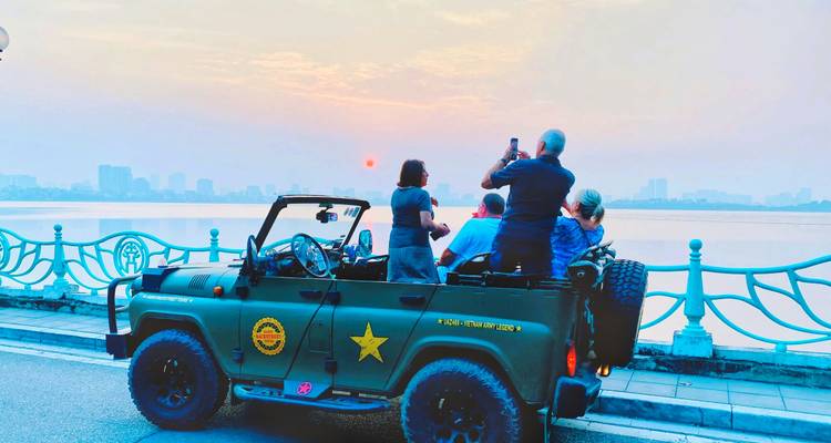 Des touristes dans une jeep militaire décapotable prennent des photos d'un lever de soleil brumeux au-dessus de l'horizon d'une ville au bord d'un lac.