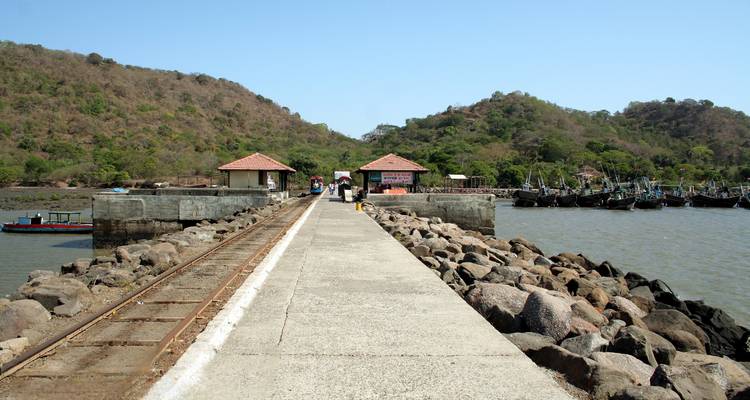 Pier, der zu kleinen Gebäuden und Booten mit bergiger Landschaft im Hintergrund führt.