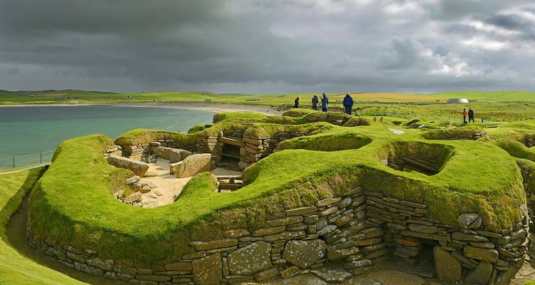 Ancient ruins of Skara Brae with a coastline view and tourists visiting.