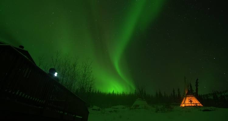 Aurora boreal sobre una terraza y tipi con una persona observando.