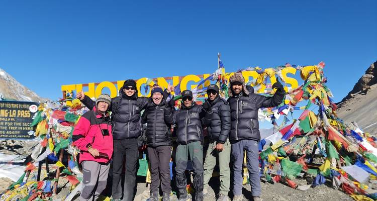 Sechs fröhliche Trekker posieren mit Gebetsfahnen beim schneebedeckten Schild des Thorong La Passes unter einem klaren Himmel.