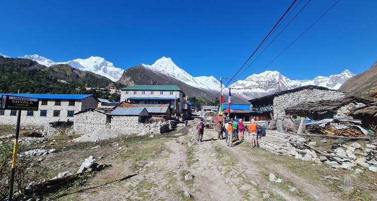 Les randonneurs parcourent un sentier rustique passant par des maisons en pierre d'un village, avec les pics enneigés de l'Himalaya en arrière-plan.