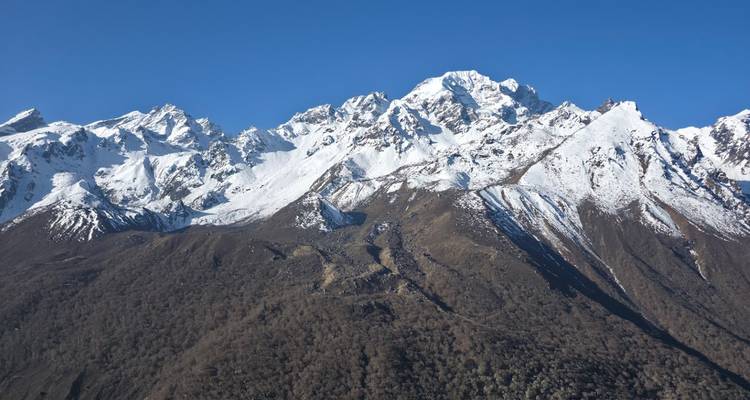 Clear view of a rugged forested valley rising to jagged snow-clad Himalayan peaks under blue sky.