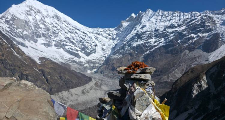 Stacked shrine stones draped with prayer flags overlooking dramatic snowy ridge.