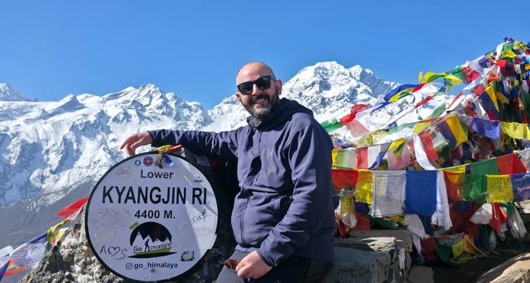 Smiling trekker points at a sign reading 'Lower Kyangjin Ri 4400 m' beside fluttering prayer flags and snowy peaks.