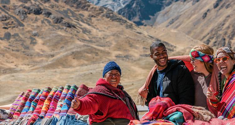 Des voyageurs partagent un rire avec un vendeur local à côté de couvertures tissées colorées sur fond de pics andins escarpés.