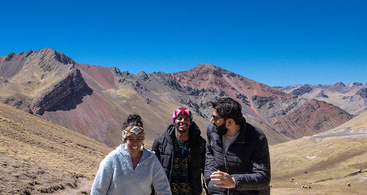 Tres amigos caminan por un sendero andino de gran altitud con montañas coloridas y un cielo azul vívido detrás de ellos.