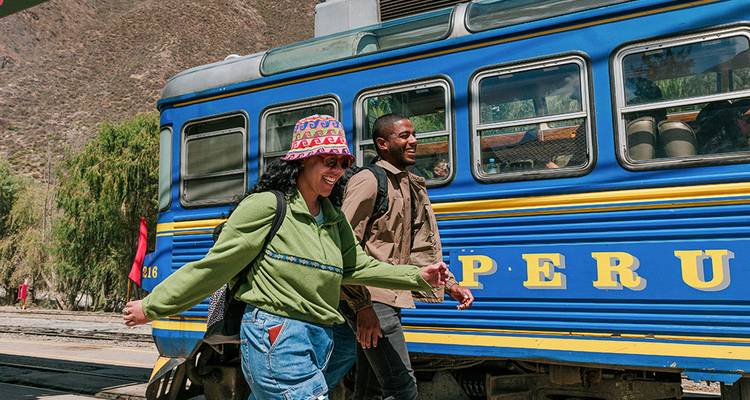 Dos viajeros sonríen mientras caminan junto a un tren PeruRail azul brillante ubicado en el Valle Sagrado.