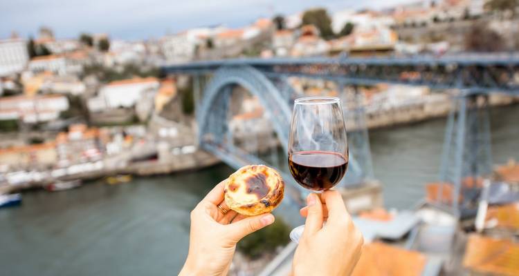 Las manos sostienen un pastel de nata y una copa de vino tinto con el Puente Dom Luís I de Oporto difuminado detrás.