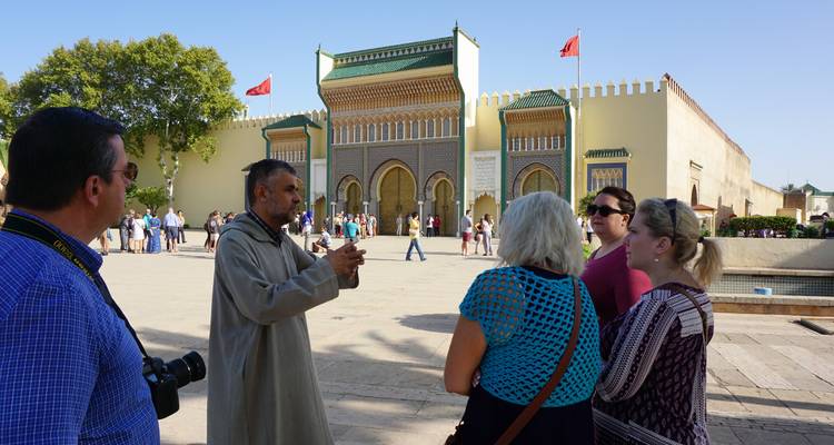 Groep toeristen luisterend naar een gids buiten een paleispoort met Marokkaanse vlaggen.