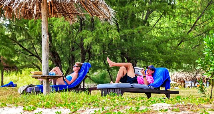 Zwei Personen entspannen auf Liegestühlen unter einem Sonnenschirm aus Stroh an einem Strand.
