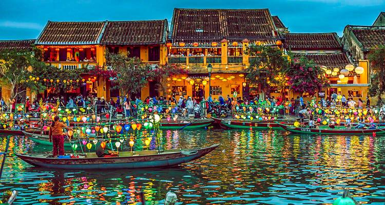 Drukke rivierscène met lantaarnverlichte boten en historische gebouwen.