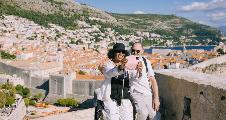Un couple souriant prenant un selfie au sommet des remparts de Dubrovnik avec les toits en terre cuite s'étendant derrière eux.