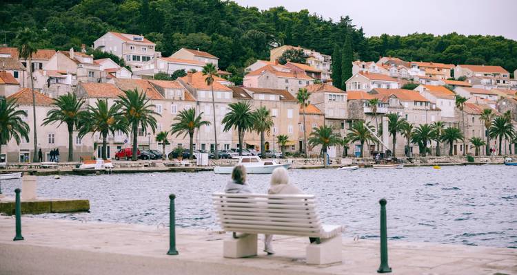 Deux voyageurs âgés sont assis sur un banc blanc face à un front de mer bordé de palmiers et à des maisons en pierre de couleur pastel.