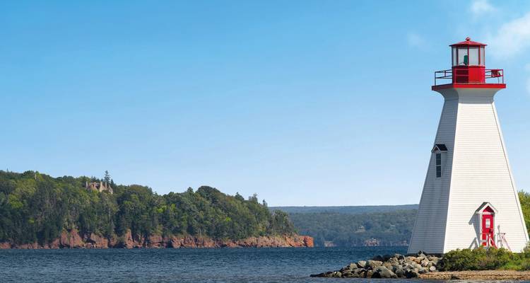 Phare blanc et rouge se dressant sur un rivage rocheux avec une eau bleue calme et des falaises boisées sous un ciel dégagé.