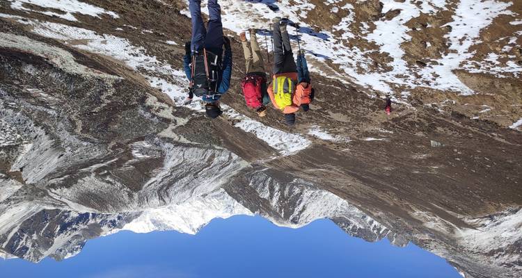 Un grupo de senderismo camina por un sendero de montaña nevado con picos escarpados por delante bajo un cielo azul despejado.