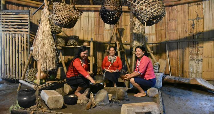 Tres mujeres indígenas preparando comida sobre fogones de piedra dentro de una choza comunal de bambú.