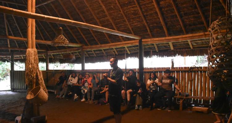 Guía hablándole a un grupo de visitantes sentados dentro de una gran casa de reuniones con techo de paja y lados abiertos.