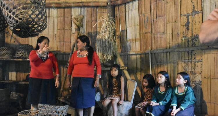 Ancianos explicando costumbres a chicas jóvenes vestidas con trajes tradicionales dentro de una casa de reuniones de bambú.