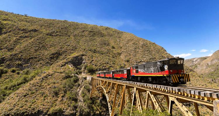 Train touristique rouge et noir traversant un pont de chevalets en bois spectaculaire à travers les flancs de collines andines escarpées et verdoyantes