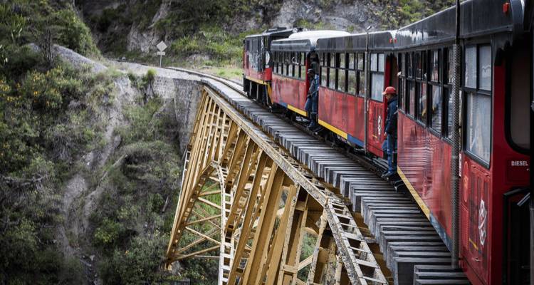 Train de passagers rouge avançant lentement sur un haut pont d'acier jaune enjambant un ravin luxuriant