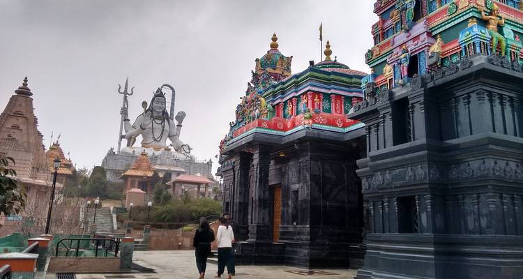 Besucher wandeln zwischen farbenfrohen südindischen Tempeln mit einer riesigen Shiva-Statue im Hintergrund.
