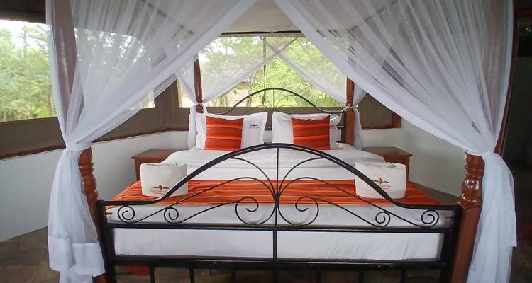Himmelbett mit weißen Moskitonetzen und orangefarbenen Akzentkissen im Inneren eines Safari-Lodge-Zeltes.