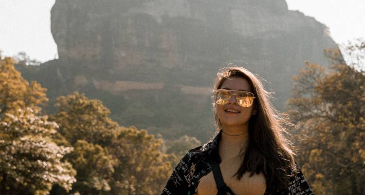 Femme souriante portant des lunettes de soleil devant la silhouette floue du Rocher de Sigiriya.
