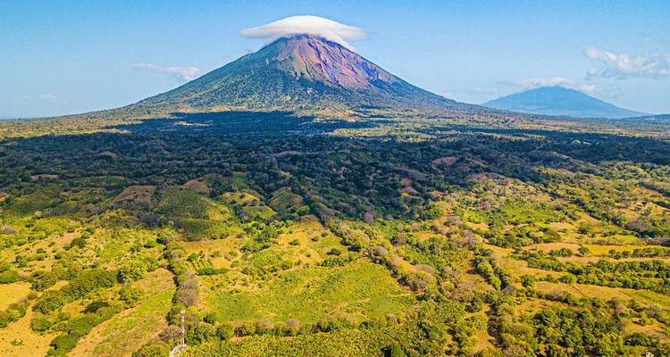 Un volcán cónico con una corona de nubes, rodeado de bosques.