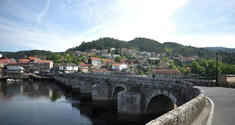Puente de piedra que lleva a un pintoresco pueblo junto al río rodeado de colinas verdes bajo un cielo despejado.