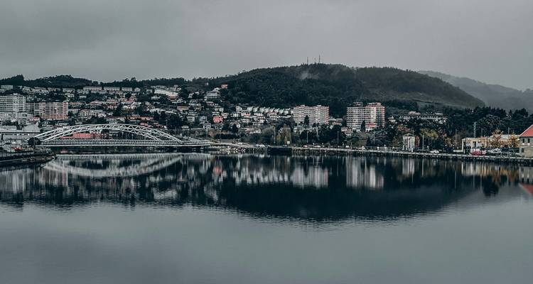Vista gris y melancólica de un puente moderno y edificios de apartamentos reflejándose en un río ancho y tranquilo bajo un cielo nublado.