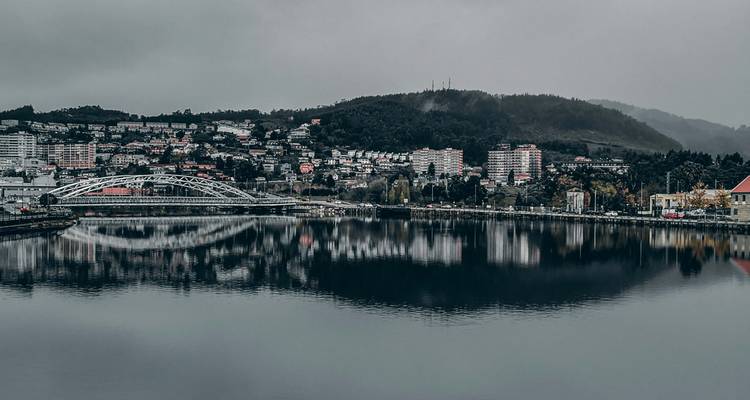 Panorama nublado de un pueblo junto al río con un puente y colinas reflejadas en las aguas oscuras y tranquilas.