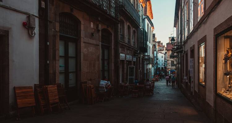 Calle estrecha y sombreada en el casco antiguo con mesas y sillas alineadas, con pocos peatones visibles a la distancia.