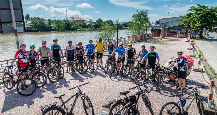 Fietsers verzamelen zich met hun fietsen op een rivierpromenade voor een groepsfoto op een zonnige dag.