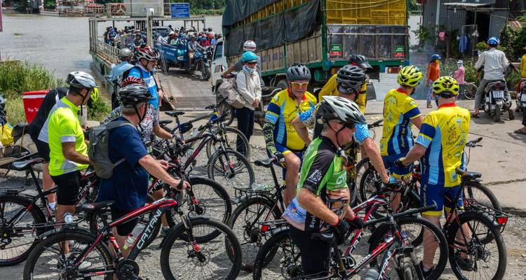 Fietsers met helmen wachten in de buurt van een veerpont over een Mekong-zijtak die druk is met lokaal verkeer.