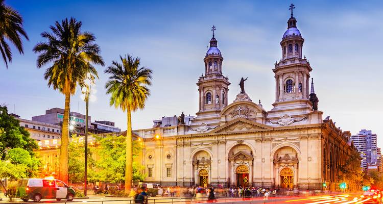 Illuminated neoclassical cathedral and palm-lined plaza bustling with evening activity in Santiago.