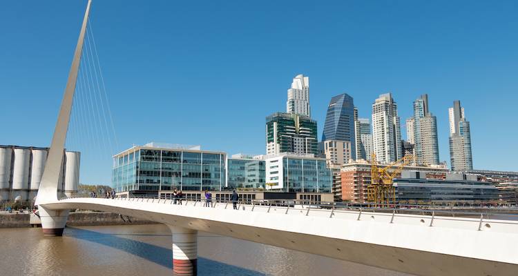 Modern bridge with sleek white pylon spanning a dockside canal against the skyline of Buenos Aires.