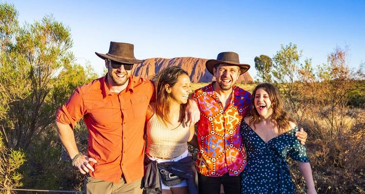 Grupo sonriente de amigos con sombreros de campo posan con Uluru brillando en rojo en el fondo.