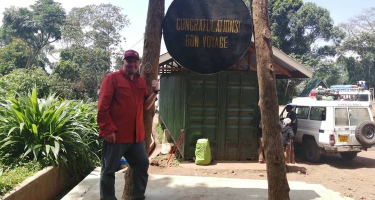 Una persona parada junto a un letrero que dice 'Congratulations Bon Voyage' con un edificio y un vehículo en el fondo.
