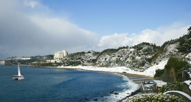 Schneebelag auf Küstenklippen und Strand von Jeju-do mit Segelboot auf tiefblauem Meer.